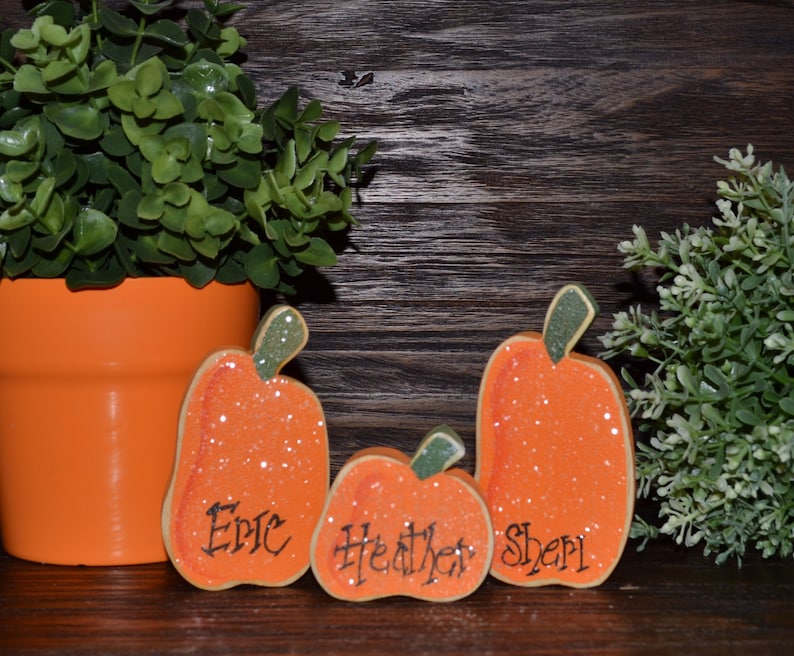 May include: Three orange wooden pumpkin decorations with green stems and glitter accents. Each pumpkin has a name written on it: Eric, Heather, and Sheri. A potted plant and greenery are in the background.
