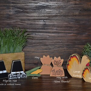 May include: A collection of wooden Thanksgiving decorations including two black pilgrim hats, one with the name Elizabeth, a yellow corn cob, two tan hay bales, one labeled Give Thanks, and two turkeys with red, yellow, and orange tail feathers.