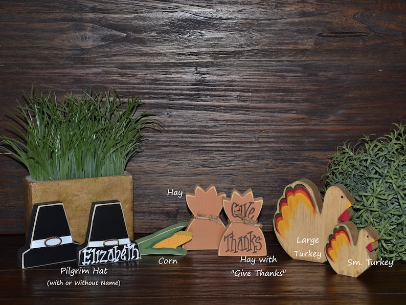 May include: A collection of wooden Thanksgiving decorations including two black pilgrim hats, one with the name Elizabeth, a yellow corn cob, two tan hay bales, one labeled Give Thanks, and two turkeys with red, yellow, and orange tail feathers.