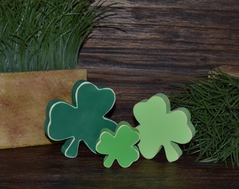 May include: Three green shamrock shapes made of wood, with a light green shamrock in the center and a darker green shamrock on either side. The shamrocks are on a brown wooden surface.
