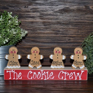 May include: Wooden gingerbread people figurines with names, standing on a red base that reads "The Cookie Crew" in white lettering. The gingerbread people have painted details and are displayed in front of a wooden background.