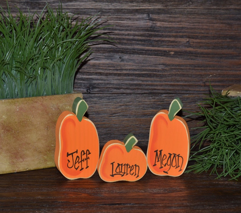 May include: Three wooden pumpkin decorations in varying sizes, painted orange with green stems. Each pumpkin has a name written on it: Jeff, Lauren, and Megan. The pumpkins are set against a rustic wooden background, suggesting a fall or harvest theme.