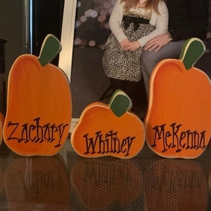 May include: Wooden pumpkin cutouts in various sizes, painted orange with green stems. Each pumpkin has a name written in black script: Yogi, Zachary, Whitney, McKenna, and Gus. A festive fall decoration.