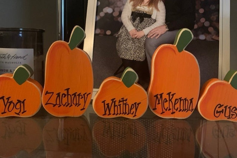 May include: Wooden pumpkin cutouts in various sizes, painted orange with green stems. Each pumpkin has a name written in black script: Yogi, Zachary, Whitney, McKenna, and Gus. A festive fall decoration.