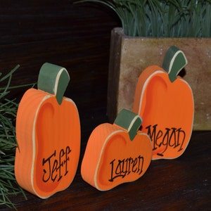 May include: Three orange wooden pumpkin decorations with green stems and the names "Jeff", "Lauren", and "Megan" in black script. The pumpkins are painted with a light orange interior and sit on a dark wooden surface.