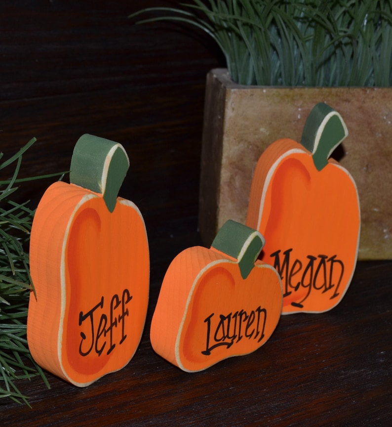 May include: Three orange wooden pumpkin decorations with green stems and the names "Jeff", "Lauren", and "Megan" in black script. The pumpkins are painted with a light orange interior and sit on a dark wooden surface.