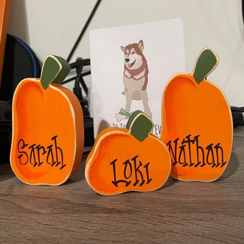 May include: Three wooden pumpkin decorations in varying sizes. Each pumpkin is painted orange with a green stem and the names "Sarah", "Loki", and "Nathan" in black script. A dog illustration is in the background.