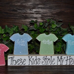 May include: A wooden sign with four glittery t-shirt cutouts in pink, blue, green, and light blue, each with a name. The sign reads "These Kids Stole My Heart."