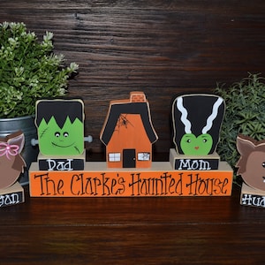 May include: A wooden Halloween display set featuring a central orange house, a green Frankenstein-style figure labeled Dad, a green bride-style figure labeled Mom, and two brown werewolf figures labeled Megan and Hudson, with the text The Clarke's Haunted House.