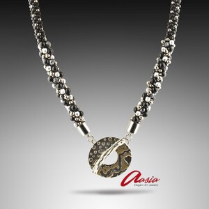 May include: A necklace with a beaded chain and a pendant. The chain is made of black and silver beads. The pendant is a silver-toned circular shape with a textured pattern and a cut-out. The necklace has the word "Aasia" in red script.