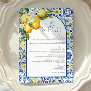 May include: A menu card with a lemon and floral design, featuring the text "The MENU". The card is set on a white plate with gold cutlery and a gold fork. The background includes white flowers and a blue fabric.