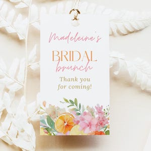 May include: A white tag with the text "Madeleine's BRIDAL brunch Thank you for coming!" in pink and orange. The tag is decorated with watercolor flowers and citrus. A gold tassel hangs from the top.