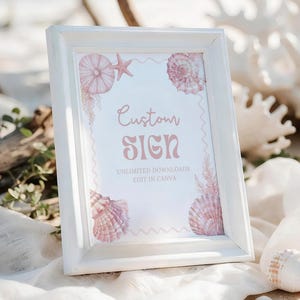 May include: A white framed sign with the words "Custom Sign" in a pink font, decorated with seashell illustrations. The sign also includes the text "Unlimited Downloads Edit in Canva". The sign is displayed on a sandy surface with seashells and coral.