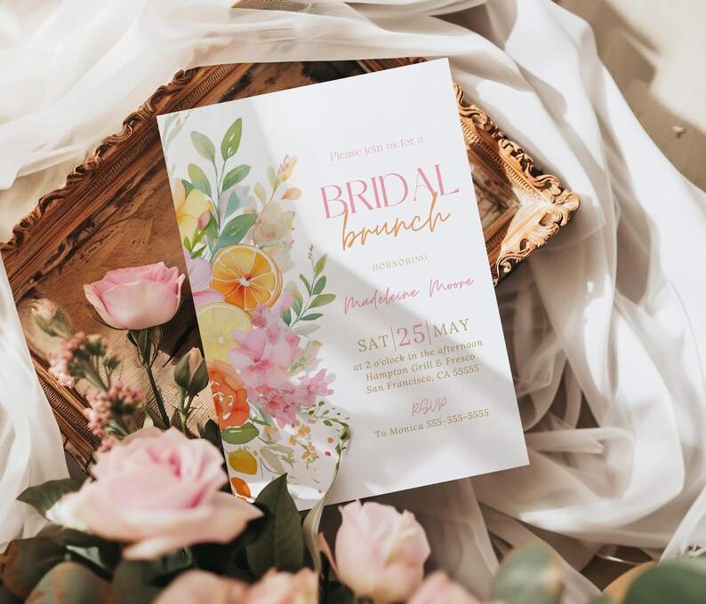 May include: A bridal brunch invitation with floral and citrus watercolor illustrations. The invitation features the text "BRIDAL brunch" and event details, including the date, time, and location. Pink roses and greenery surround the invitation, set on a wooden tray.