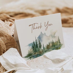 Peut inclure: Carte blanche pliée avec l'inscription "Thank You" en écriture brune. La carte présente un paysage à l'aquarelle d'une chaîne de montagnes et de conifères verts et bleus.