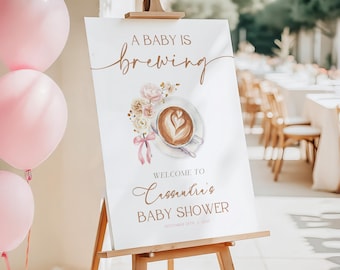 May include: A white sign on a wooden easel with the text "A BABY IS BREWING" in elegant script. A watercolor illustration of a coffee cup with a heart-shaped design and floral accents is featured. The sign welcomes guests to a baby shower.