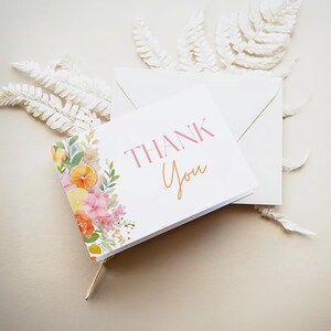 May include: A white card with the words "THANK You" in pink and orange, with a floral design of lemons and pink flowers. The card is next to a white envelope and white decorative leaves.