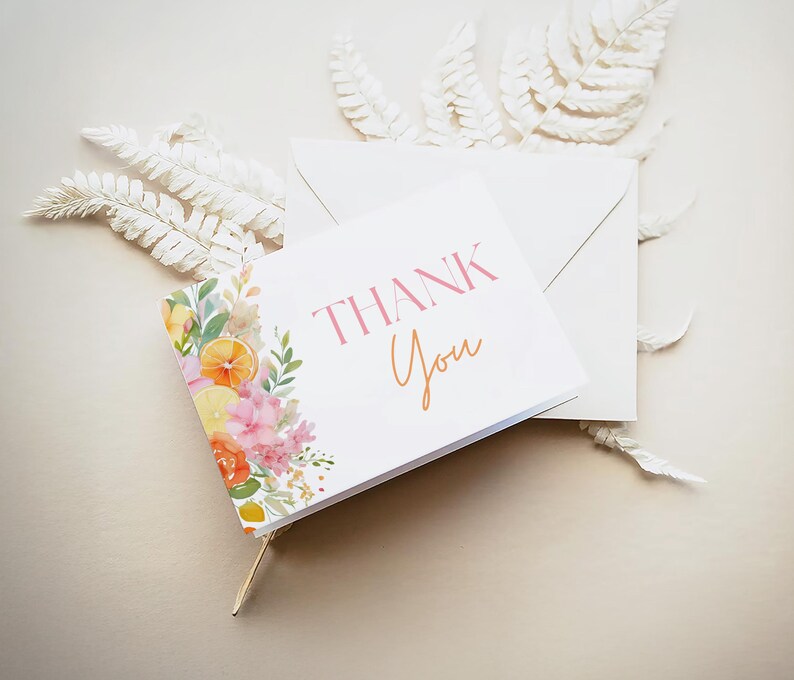 May include: A white card with the words "THANK You" in pink and orange, with a floral design of lemons and pink flowers. The card is next to a white envelope and white decorative leaves.