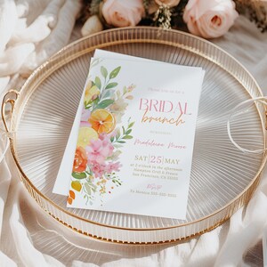 May include: A white invitation for a bridal brunch, featuring floral and citrus watercolor illustrations. The invitation is on a gold-rimmed tray with the text "BRIDAL brunch" and event details. Ribbons and flowers surround the tray.