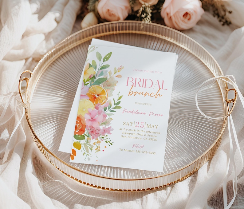 May include: A white invitation for a bridal brunch, featuring floral and citrus watercolor illustrations. The invitation is on a gold-rimmed tray with the text "BRIDAL brunch" and event details. Ribbons and flowers surround the tray.