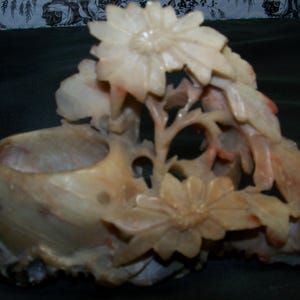 May include: A carved stone floral sculpture featuring a bowl-like base with intricate details. The sculpture showcases blooming flowers and leaves in shades of beige and light orange, creating a naturalistic design.