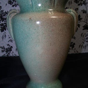 May include: A tall, vintage ceramic vase with a two-tone color scheme. The upper portion and flared rim are a soft teal, transitioning to a tan hue in the middle, and a teal base. The vase has two handles and a speckled texture.