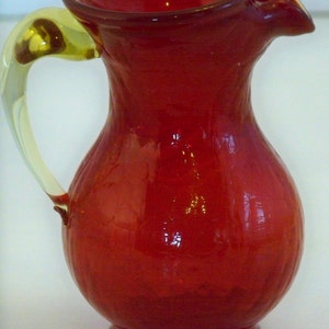 May include: A red glass pitcher with a yellow glass handle. The pitcher has a rounded body and a flared lip.