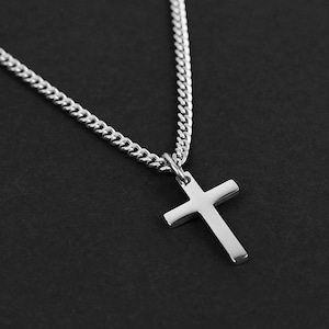 May include: A silver cross pendant necklace. The cross is a simple, minimalist design, suspended from a delicate chain. The necklace is displayed against a dark background, highlighting the jewelry's metallic sheen.