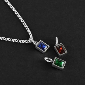 May include: A silver necklace with a chain and three rectangular pendants. Each pendant features a different colored gemstone: blue, red, and green. The pendants are set in a silver frame, and the necklace is displayed against a dark background.