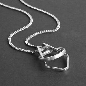 May include: A silver-toned necklace with a geometric pendant. The pendant features a ring, a triangular shape, and a five-sided shape. The chain is a curb style. The background is a dark gray color. Text at the bottom reads "Ring not included."