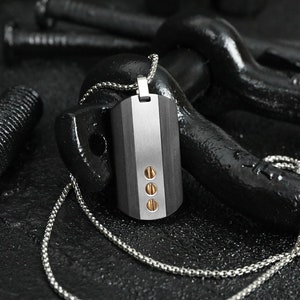 Könnte beinhalten: Eine silberne Kettenkette mit einem schwarzen und silbernen Hundemarke-Anhänger. Der Anhänger hat drei goldene Akzente.