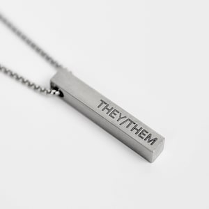 May include: A silver chain necklace with a silver bar pendant that says "THEY/THEM" on it.