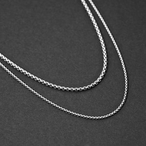 May include: Two silver chain necklaces layered on a black background. The chains are both thin and delicate.
