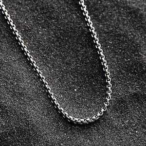 May include: A silver-toned metal chain necklace featuring a box link design. The chain is composed of small, square-shaped interlocking links that create a smooth, continuous, and flexible strand with a polished, reflective metallic finish.