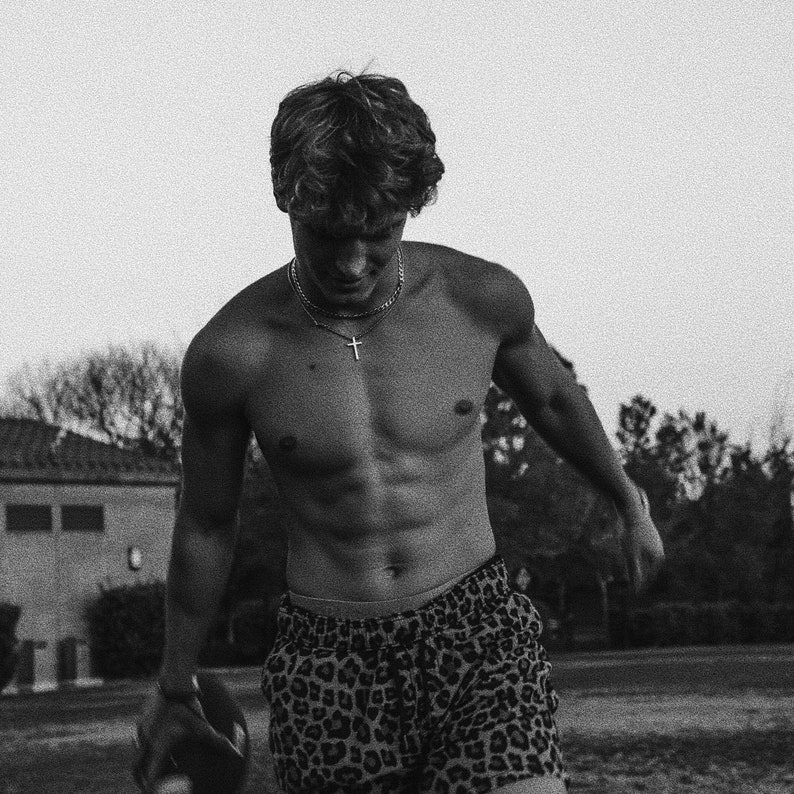 May include: A black and white photo of a person wearing leopard print shorts and a silver chain with a cross pendant.