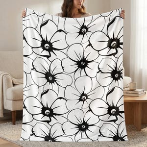 May include: A white blanket with a large black floral pattern. The flowers have a detailed outline and a dark center. The blanket is held up, showcasing the design. The background includes a beige sofa and a wooden table.