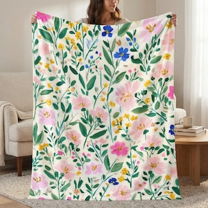 May include: A floral blanket featuring a cream background with a pattern of pink, yellow, and blue flowers and green leaves. The blanket is held up, showcasing the full design. The flowers are in various sizes and shapes, creating a vibrant and cheerful aesthetic.