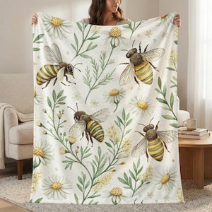 May include: A white blanket featuring a watercolor pattern of bees, daisies, and green foliage. The bees have yellow and black stripes. The daisies have white petals and yellow centers. The blanket is held up by a person.