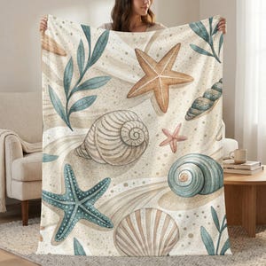 May include: A beige blanket featuring a watercolor design of seashells, starfish, and seaweed. The blanket has a sand-like background with light brown and blue accents. The design includes various sizes and colors of starfish and seashells.