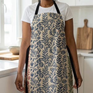 May include: Apron with a beige background and a blue floral pattern. The apron has black straps and a bib design. The floral pattern includes leaves and flowers. The apron is made of a durable fabric, suitable for kitchen use.