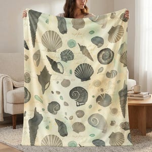 May include: A beige blanket featuring a watercolor design of various seashells in shades of gray and brown. The blanket has a vintage aesthetic with subtle script-like text scattered across the surface. The shells vary in size and shape, creating a coastal theme.