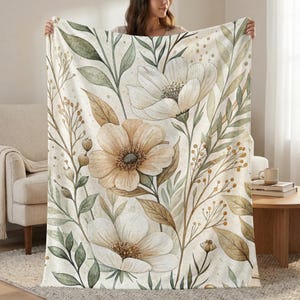 May include: A cream-colored blanket featuring a watercolor floral design. The design includes large, stylized flowers in shades of white and tan, with green and brown leaves and stems. The blanket is held up, showcasing the full pattern.