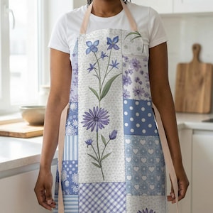 May include: A patchwork apron featuring a floral design with purple and blue flowers. The apron has a light beige neck strap and is composed of various patterned squares, including polka dots, stripes, and hearts. The apron is worn by a person in a white t-shirt.