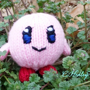 May include: Pink knitted plush toy with a smiling face, blue eyes, and red shoes. The toy is sitting in a bed of green plants.
