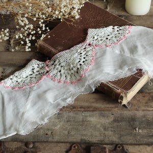 May include: A vintage white fabric with a delicate lace trim, accented with pink edging, rests atop a weathered, antique book. The fabric's edges are frayed, adding to its antique charm. The scene is set on a rustic wooden surface.