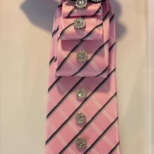 May include: Pink tie with a diagonal striped pattern in black and white. The tie is adorned with sparkling rhinestone embellishments and a large black and silver flower at the knot. A stylish accessory.