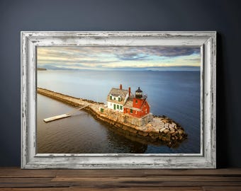 May include: A framed print of a lighthouse on a rocky island, connected to the mainland by a stone pier. The lighthouse is red with a black lantern, and the sky is cloudy with a hint of blue.