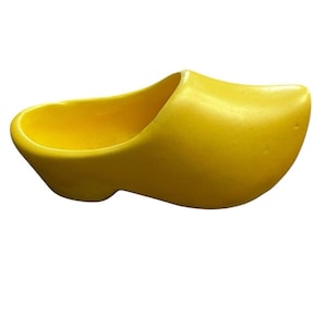 Puede incluir: Un zueco de madera amarillo brillante, un zapato tradicional holandés, se muestra sobre un fondo blanco. El zapato tiene una punta redondeada y un interior ahuecado. El zueco está hecho de un material liso y sólido.