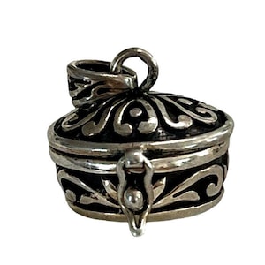 Puede incluir: Un colgante de plata en forma de medallón con un diseño ornamentado. El medallón tiene una tapa con bisagras y un pequeño pestillo. El medallón tiene forma ovalada y tiene un fondo negro con filigranas plateadas.