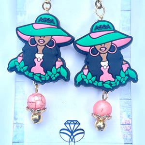 May include: A pair of dangle earrings featuring a rubberized charm of a person wearing a green and pink wide-brimmed hat with the text AKA, long dark hair, and pink hoop earrings. Below the charm hangs a pink crackle bead and a small gold bead.